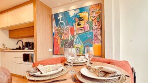 Un coin repas avec une table ronde en bois, dressée pour deux personnes, agrémentée de sets de table en fibres tressées et de couverts. Un tableau abstrait aux couleurs vives orne le mur derrière la table.