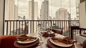 Un coin repas avec une table dressée pour deux personnes, une vue sur de hauts immeubles à travers de grandes fenêtres et une balustrade de balcon.