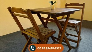 A dining area with a wooden table and chairs, a small vase with yellow flowers, a mounted TV, and a window with a view of greenery. - Chalé Pico Alto (Guaramiranga)