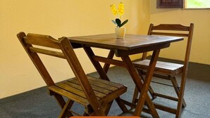 Uma área de refeições com mesa e cadeiras de madeira, um pequeno vaso com flores amarelas, TV na parede e janela com vista para vegetação.