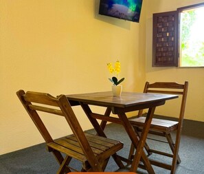 Uma área de refeições com mesa e cadeiras de madeira, um pequeno vaso com flores amarelas, TV na parede e janela com vista para vegetação.