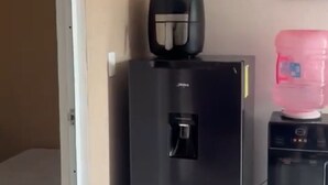 Uma geladeira compacta com dispensador de água, um pequeno alto-falante preto e um dispensador de água rosa em cima.
