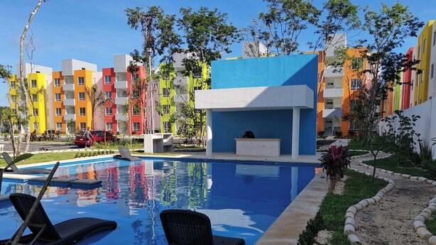 Piscina com espreguiçadeiras, cercada por edifícios coloridos e vegetação.