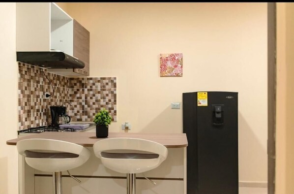 A compact kitchen with a tiled backsplash, a black refrigerator, two white bar stools, and a small potted plant. - Hostal la terraza sm (Santa Marta)