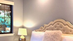 A large bed with a tufted headboard, a bedside lamp, and a window with a view of trees.