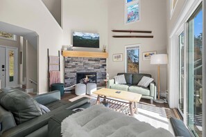 A spacious living room with a stone fireplace, a large flat-screen TV, a green sofa, and a wooden coffee table. - Modern Chalet Across from Holiday Valley – Ski-In Comfort & Year-Round Charm (Ellicottville)