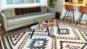 A living room with a sofa, a coffee table with plants, a patterned rug, and a kitchen area with wooden cabinets and a bar stool. - Garden of the Gods Retreat - Upper Unit (Colorado Springs)
