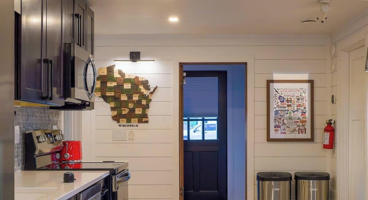 A kitchen with dark cabinets, a red appliance, a wall-mounted map of Wisconsin, a door, and two trash bins.