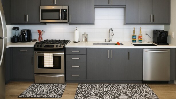 A modern kitchen with dark grey cabinets, a stainless steel microwave, a gas stove, a dishwasher, and a sink. There are two patterned rugs on the floor.
