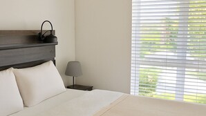 A neatly made bed with a headboard, a bedside lamp, and a window with blinds.