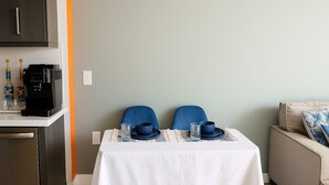 A compact kitchen with a coffee machine, a dining table set with blue chairs, and a sofa with a cushion.