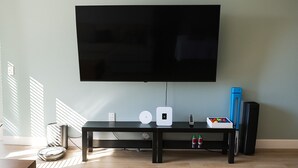 A modern living room with a flat-screen TV mounted on the wall, a black entertainment unit with speakers, a clock, and a small table with a box of colorful items.