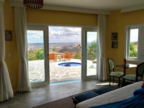 Un dormitorio con cama, una mesa, sillas y vistas a una piscina y paisaje a través de un ventanal corredero.