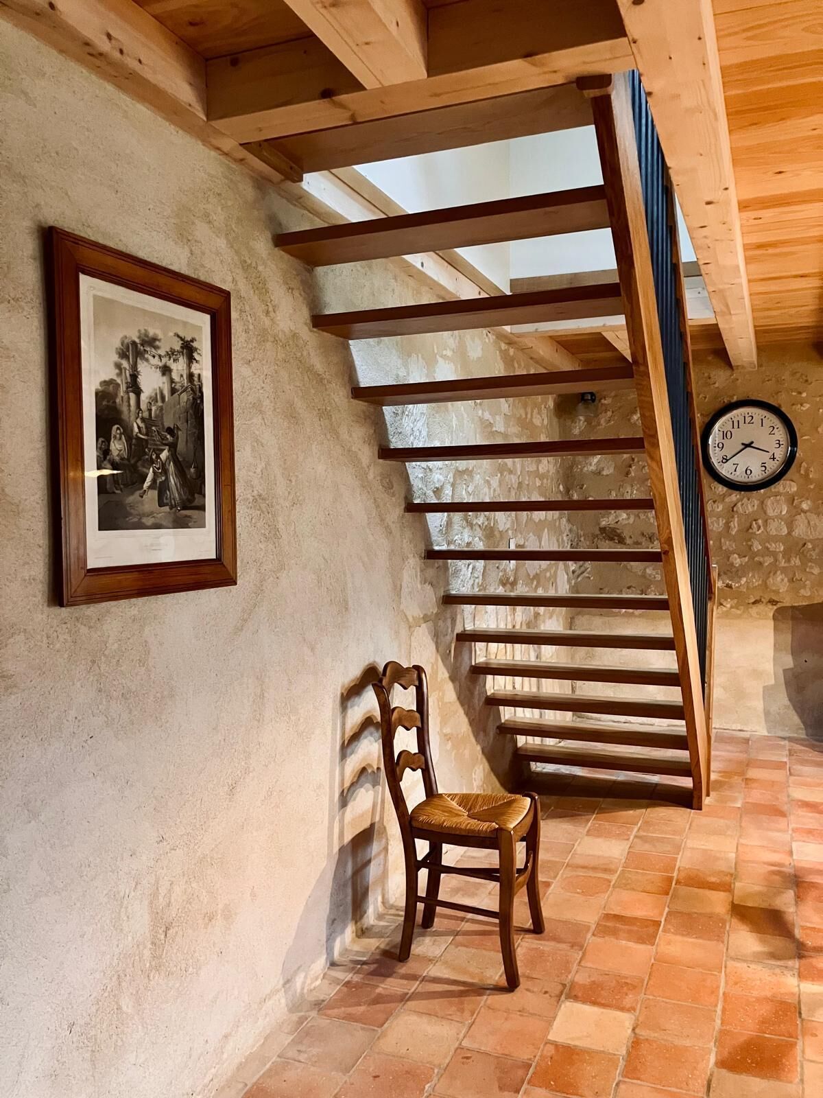 A rustic interior with a wooden staircase, a framed historical painting, a wooden chair, and a wall clock.