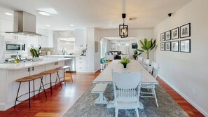 A modern kitchen with a central island, wooden stools, and a dining area with a long table and white chairs. - The Clubhouse by Coastline Vacation Rentals (San Diego)