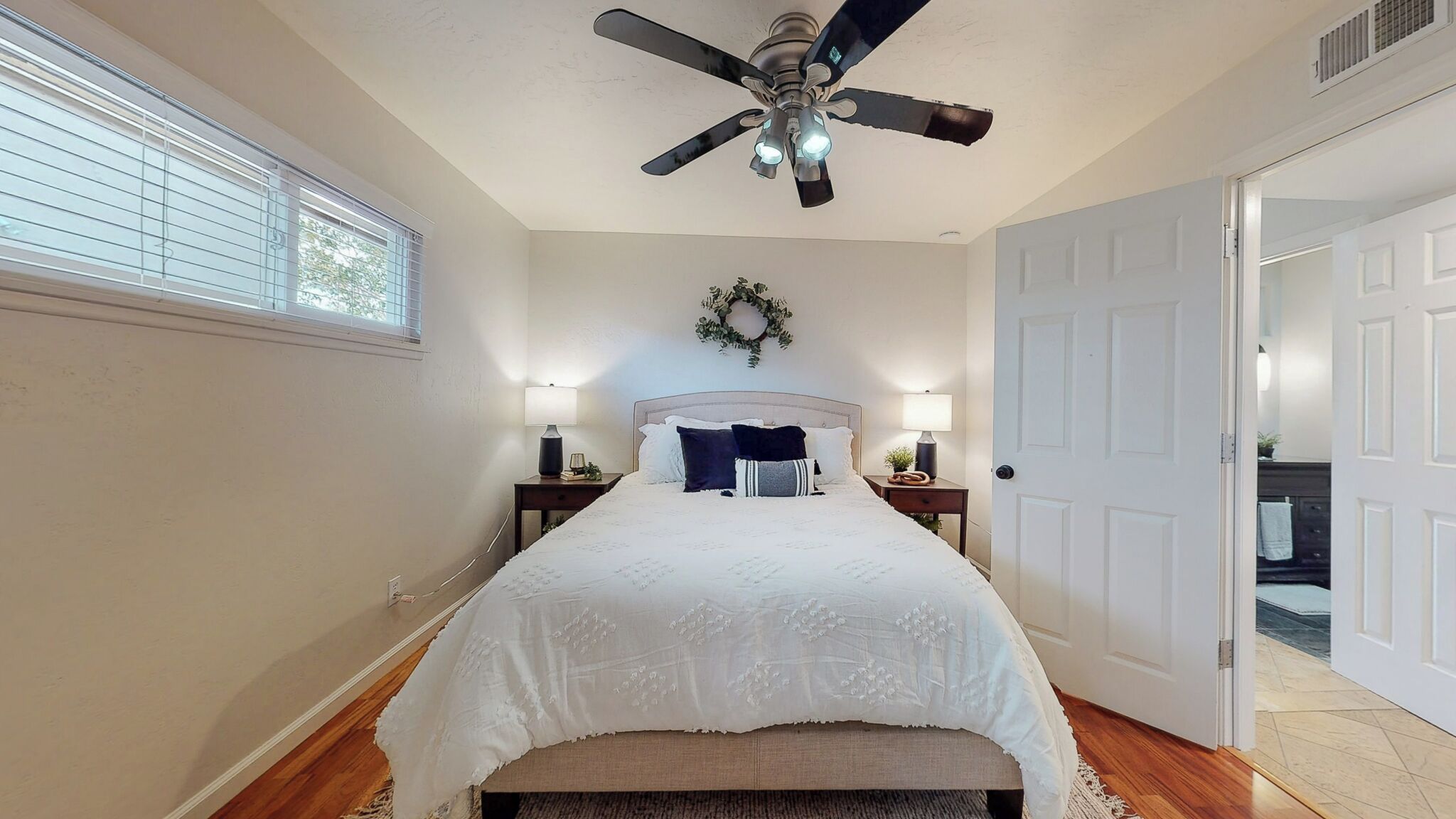 Une chambre à coucher comprenant un lit, deux tables de chevet, un ventilateur de plafond, une fenêtre avec des stores et une porte donnant sur une autre pièce.