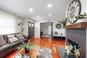 A modern living room with a dark leather sofa, a coffee table, and a fireplace. There is a large clock on the wall and a door leading to another room. - The Clubhouse by Coastline Vacation Rentals (San Diego)