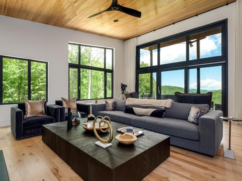 En moderne stue med træloft, store vinduer, en grå sofa, sofabord og udsigt til grøn natur.