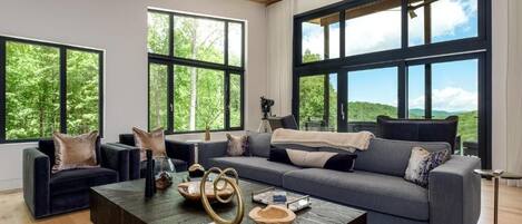 En moderne stue med træloft, store vinduer, en grå sofa, sofabord og udsigt til grøn natur.