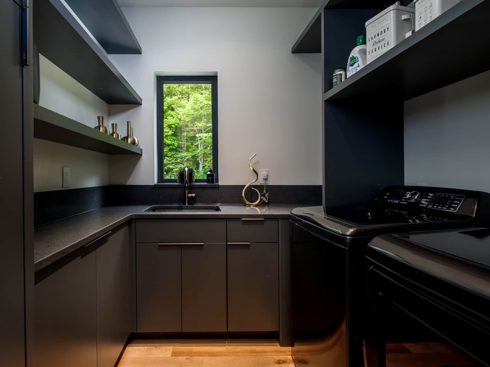 A modern kitchen with a built-in stove, a window with a view of greenery, and a shelf with a laundry service sign.