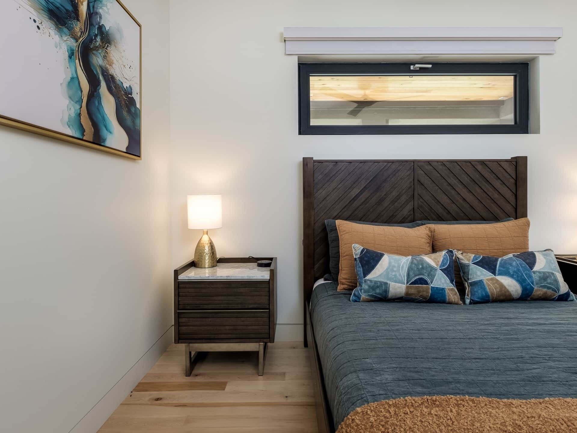 A bedroom with a bed, a nightstand with a lamp, a window above the bed, and a wall-mounted shelf.