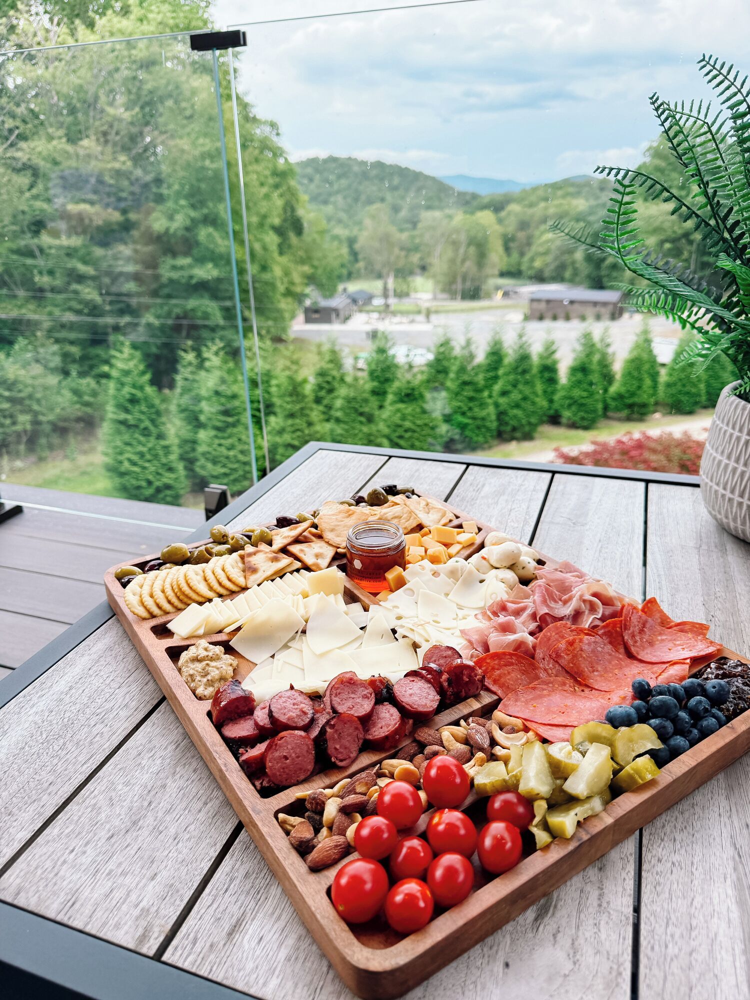 A wooden tray with various cheeses, meats, and fruits on a balcony with a view of a forest and a building.