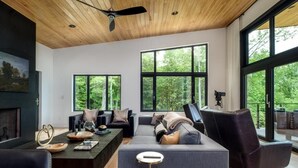 A modern living room with a large sectional sofa, a coffee table, and a fireplace. There are large windows providing a view of the outdoors. - Luxury Home - The Retreat House (Balsam Grove)