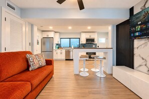 A modern kitchen with white cabinets, a stainless steel refrigerator, and a built-in oven. There is a bar area with stools and a TV mounted on the wall displaying a movie. - Clearwater Bliss Apartments 3 (Clearwater)