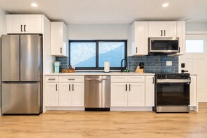 A modern kitchen with stainless steel appliances, white cabinetry, and a large window. - Clearwater Bliss Apartments 3 (Clearwater)
