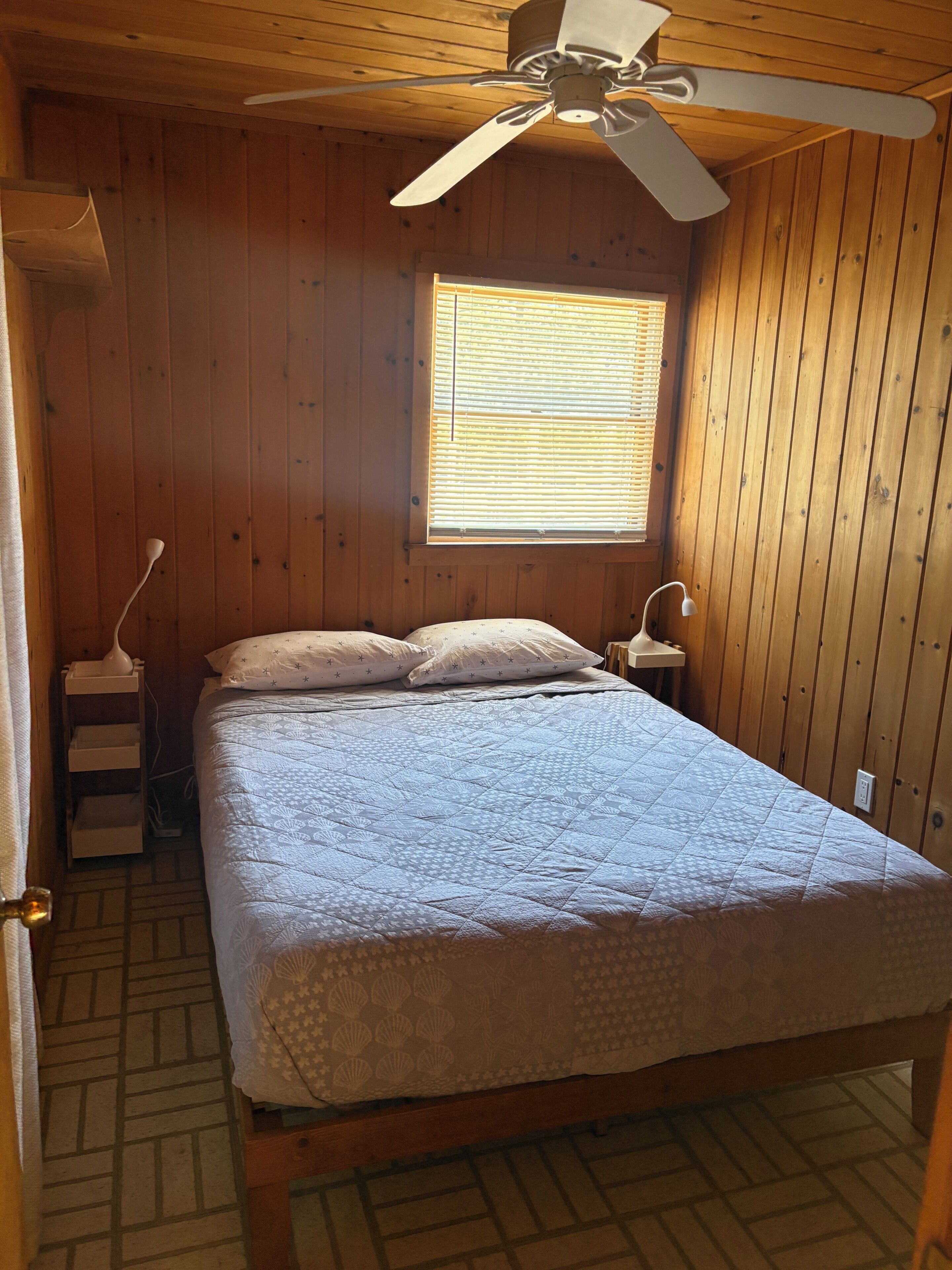 Una habitación con paneles de madera, una cama, un ventilador de techo, una ventana con persianas y una mesita de noche con lámpara.