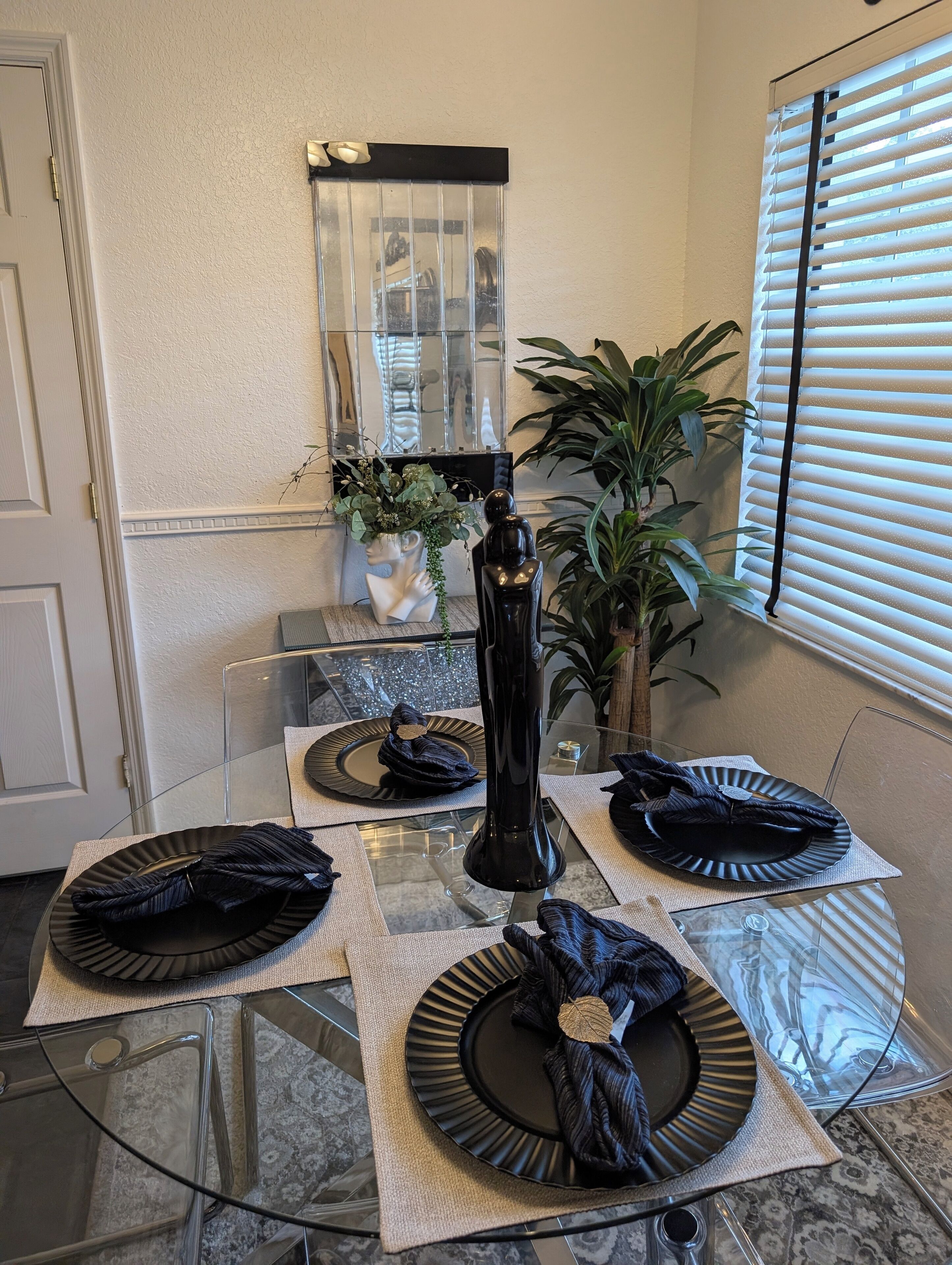Un coin repas avec une table en verre dressée pour quatre personnes, agrémentée d’assiettes noires et d’un grand vase noir.