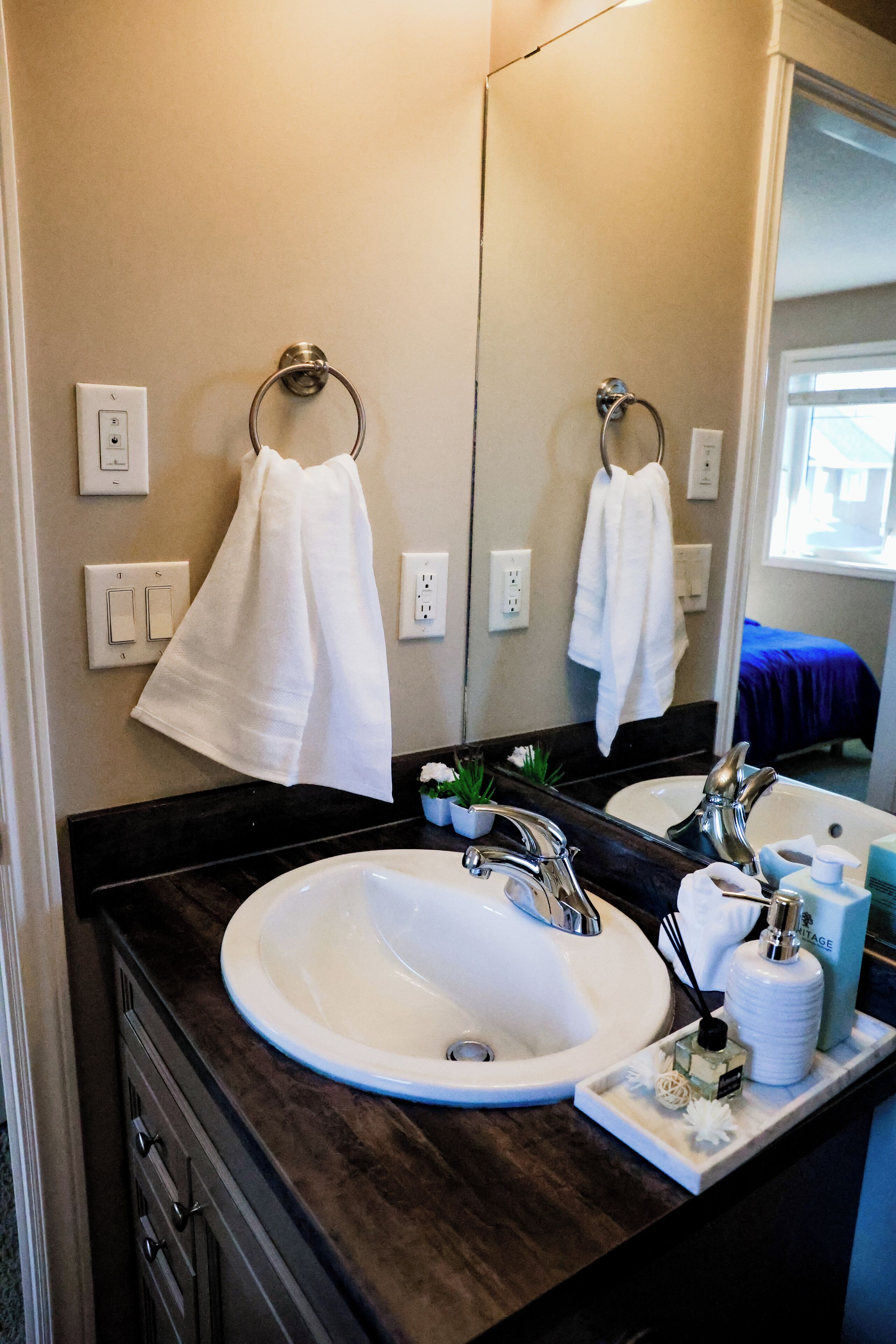 A bathroom with a white sink, dark countertop, and a mirror reflecting a bed with a blue blanket.