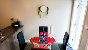 Un coin repas avec une table en verre, des chaises noires, une horloge au mur et un comptoir de cuisine équipé d’appareils électroménagers.