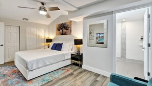 Un dormitorio moderno con cama, ventilador de techo, un cuadro, mesita de noche y baño visible a través de una puerta abierta.