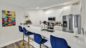 Una cocina moderna con armarios blancos, electrodomésticos de acero inoxidable y encimera de mármol. Hay tres taburetes azules y un adorno en la pared.
