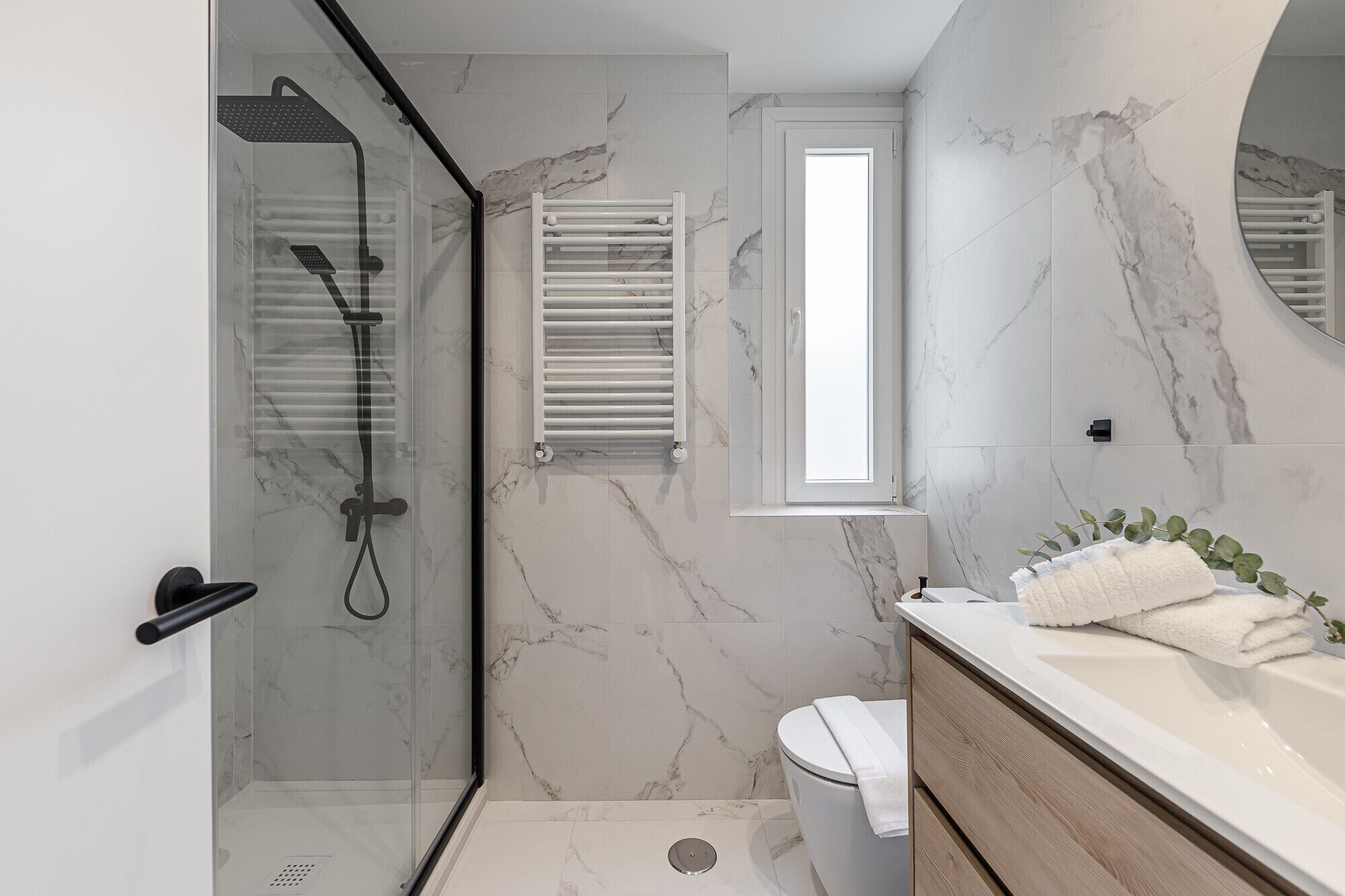 Une salle de bain moderne avec une douche à parois de verre, un meuble-lavabo blanc avec un miroir et un mur recouvert de carreaux de marbre.