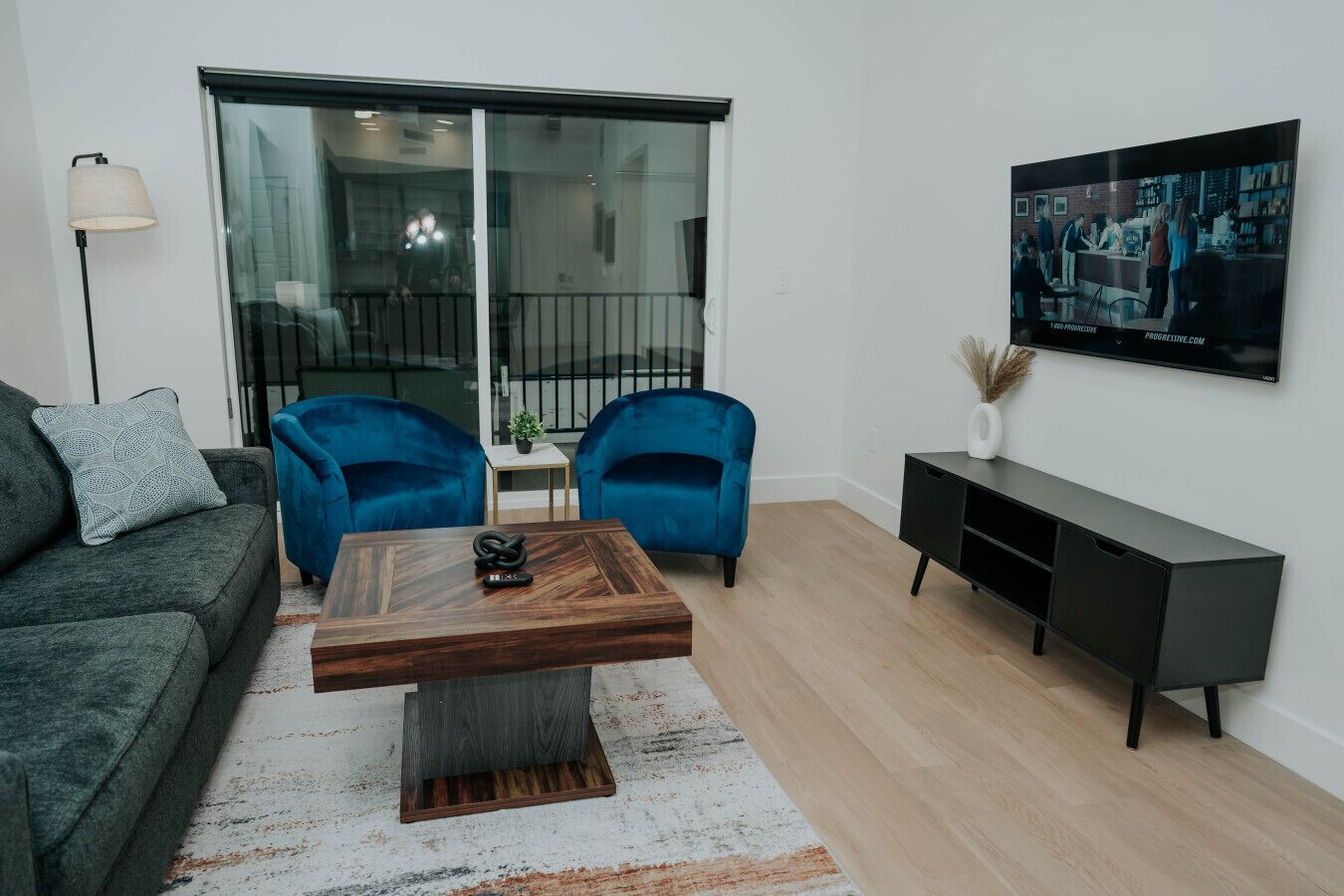 A modern living room with a grey sofa, two blue armchairs, a wooden coffee table, a black TV stand, and a large window with a view of the outside.