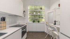 A compact kitchen with white cabinetry, a built-in oven, a window with a tropical wallpaper, and a coffee machine. - Waou Sol VII - Short Term Rental (Madrid)