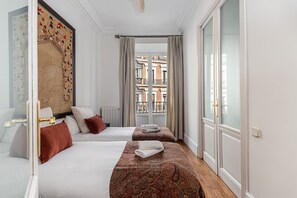 Une chambre d’hôtel avec deux lits, une grande fenêtre donnant sur des immeubles et une tapisserie à motifs accrochée au mur.