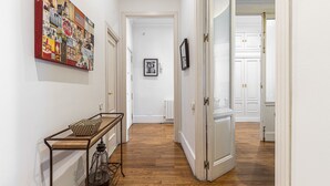 A hallway with a wooden floor, a metal bench with a checkered cushion, a wall-mounted painting, and a white door with a brass handle. - Waou Sol VII - Short Term Rental (Madrid)