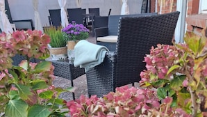 An outdoor seating area with wicker chairs and a table, surrounded by potted plants and flowers.