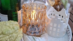 A table with a green bottle, two clear glasses, a lit candle, a white decorative house, and a basket.