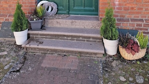 A green double door with a sign above it, a brick wall, and two potted plants on the steps.