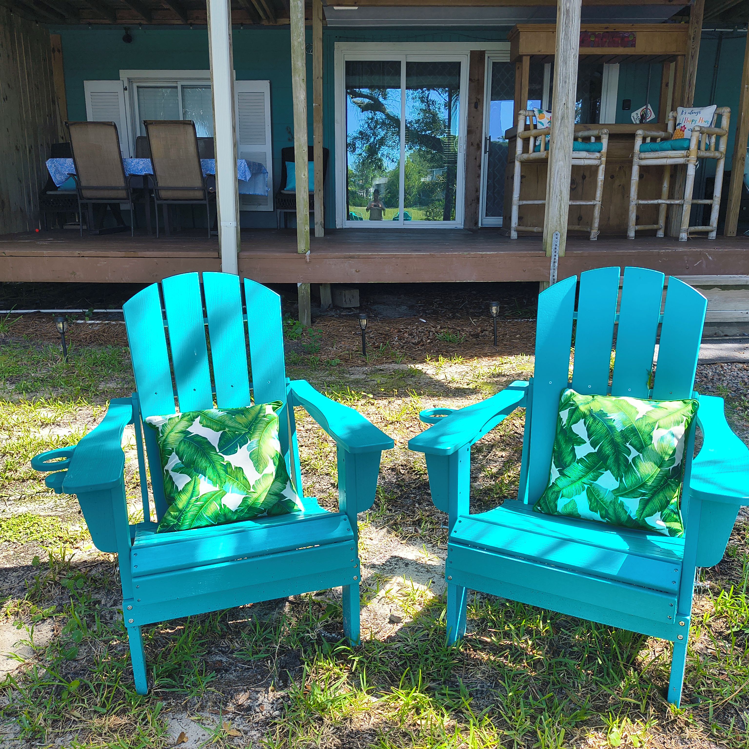 Zwei türkisfarbene Adirondack-Stühle mit Kokosblattkissen stehen auf einer grünen Fläche vor einer Holzdiele.