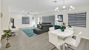 Uma sala de estar moderna com um sofá seccional cinza, uma mesa de jantar branca com quatro cadeiras, um tapete azul estampado e um vaso com planta.