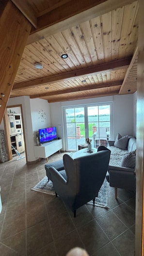 Ein Wohnzimmer mit Fernseher, Sofa und Blick auf ein Gewässer durch große Fenster.