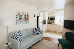 Ein modernes Wohnzimmer mit einem hellblauen Sofa, gestreiften Kissen und einer Stehlampe. Ein grüner Sessel ist im Vordergrund zu sehen. Das Zimmer geht in eine Küche mit weißen Schränken und einem Edelstahlkühlschrank über.