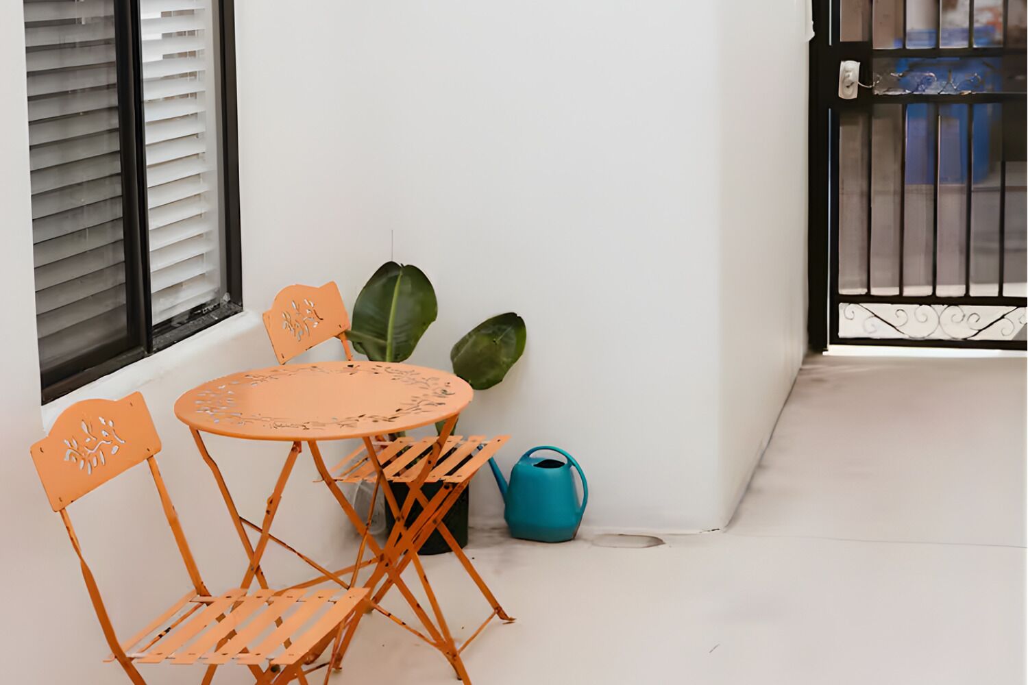 Uma pequena área externa com mesa e cadeiras, uma planta em vaso e um regador azul.