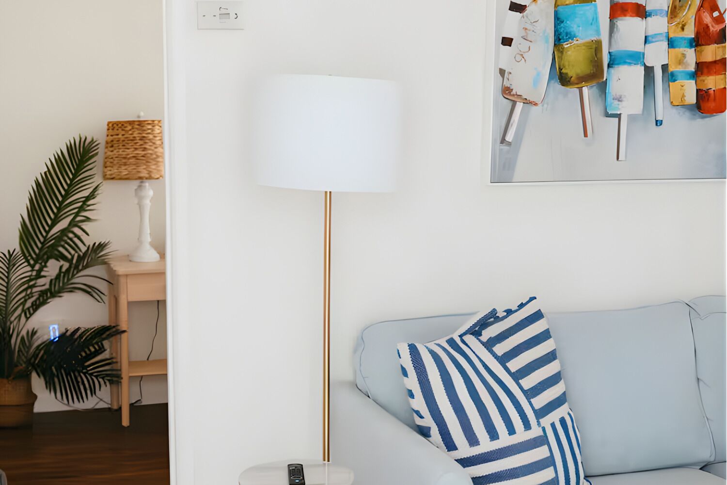 Ein Wohnzimmer mit einem hellblauen Sofa, einem gestreiften Kissen, einer Stehlampe, einem kleinen Tisch mit Lampe und einem Wandbild.