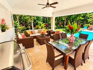 Uma área de estar coberta ao ar livre, com mobiliário de vime, uma mesa de vidro e vista para uma piscina e vegetação exuberante.